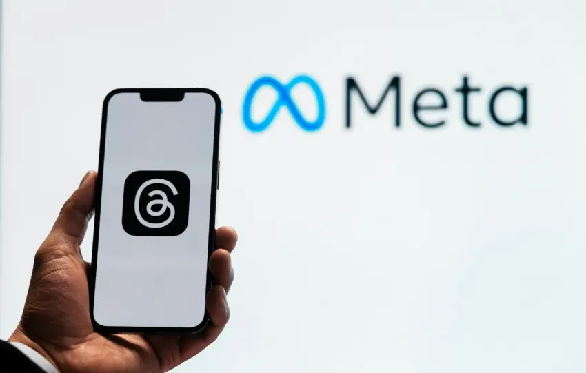 A hand holding a smartphone showing the Threads app with Meta logo in the background.
